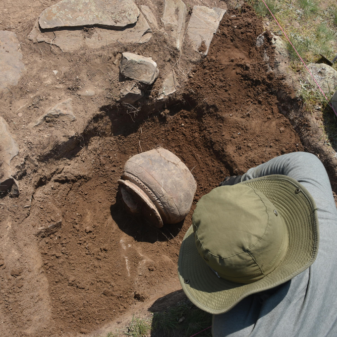 A person wearing a green hat bends over a dug hole in which a piece of pottery rests.